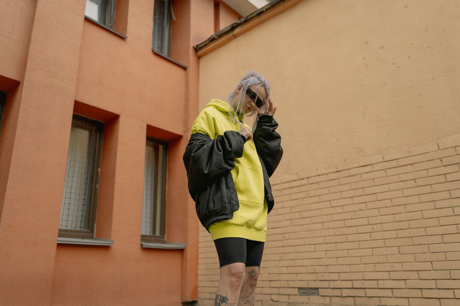 Trendy woman in a yellow hoodie and sunglasses posing outdoors by a building wall.
