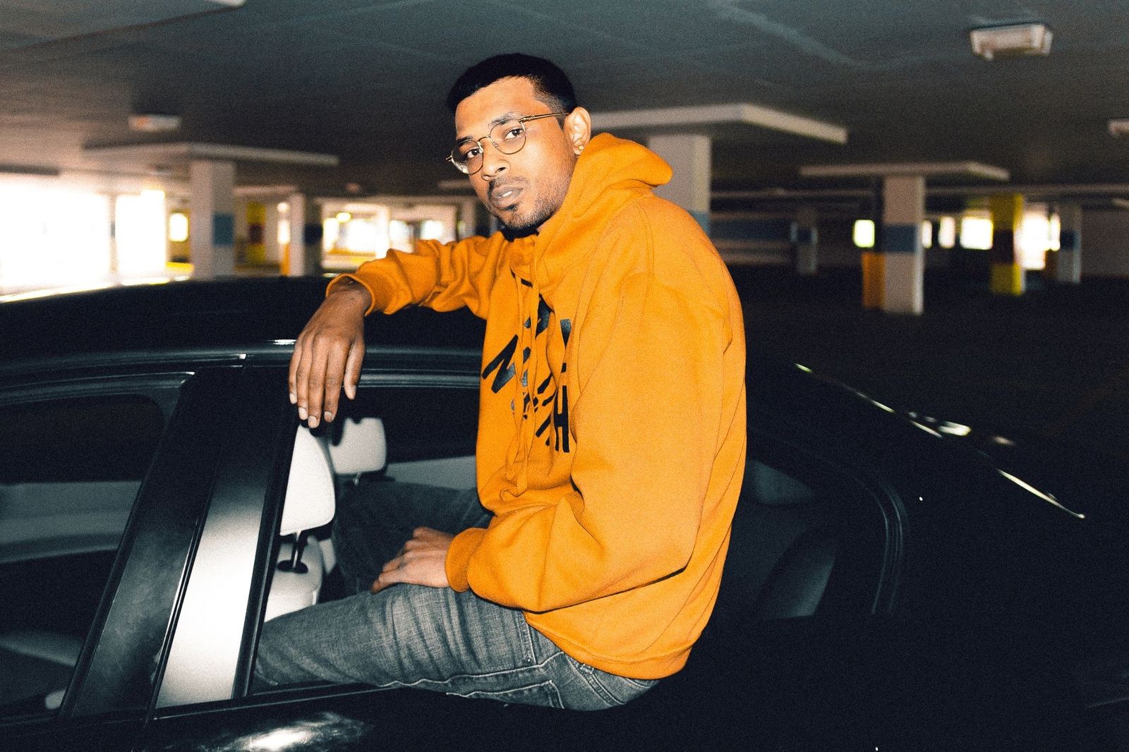 Casual man in an orange hoodie sits on a car in a dimly lit parking garage.