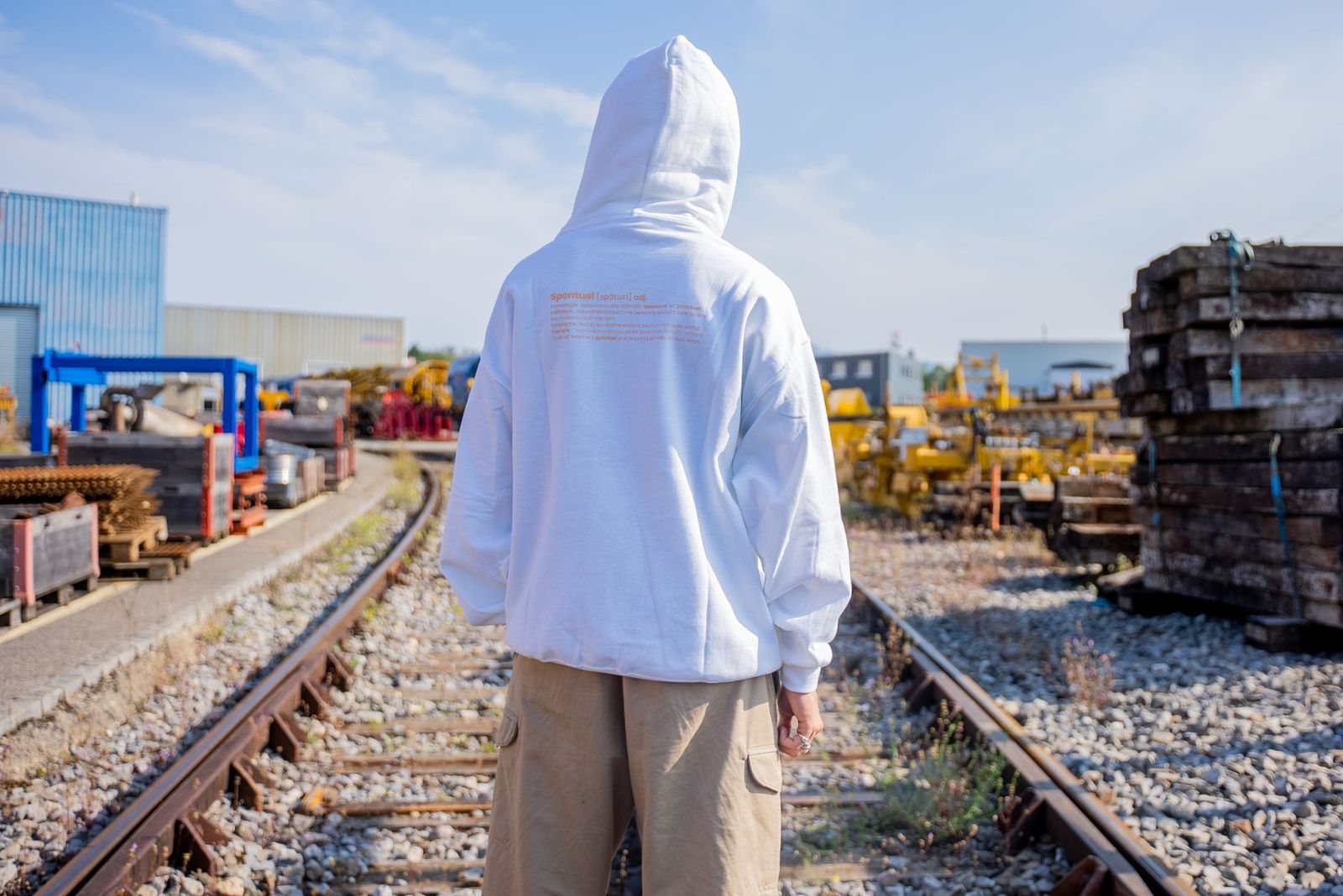 Back view of a person in a white hoodie standing on railway tracks in an industrial setting.